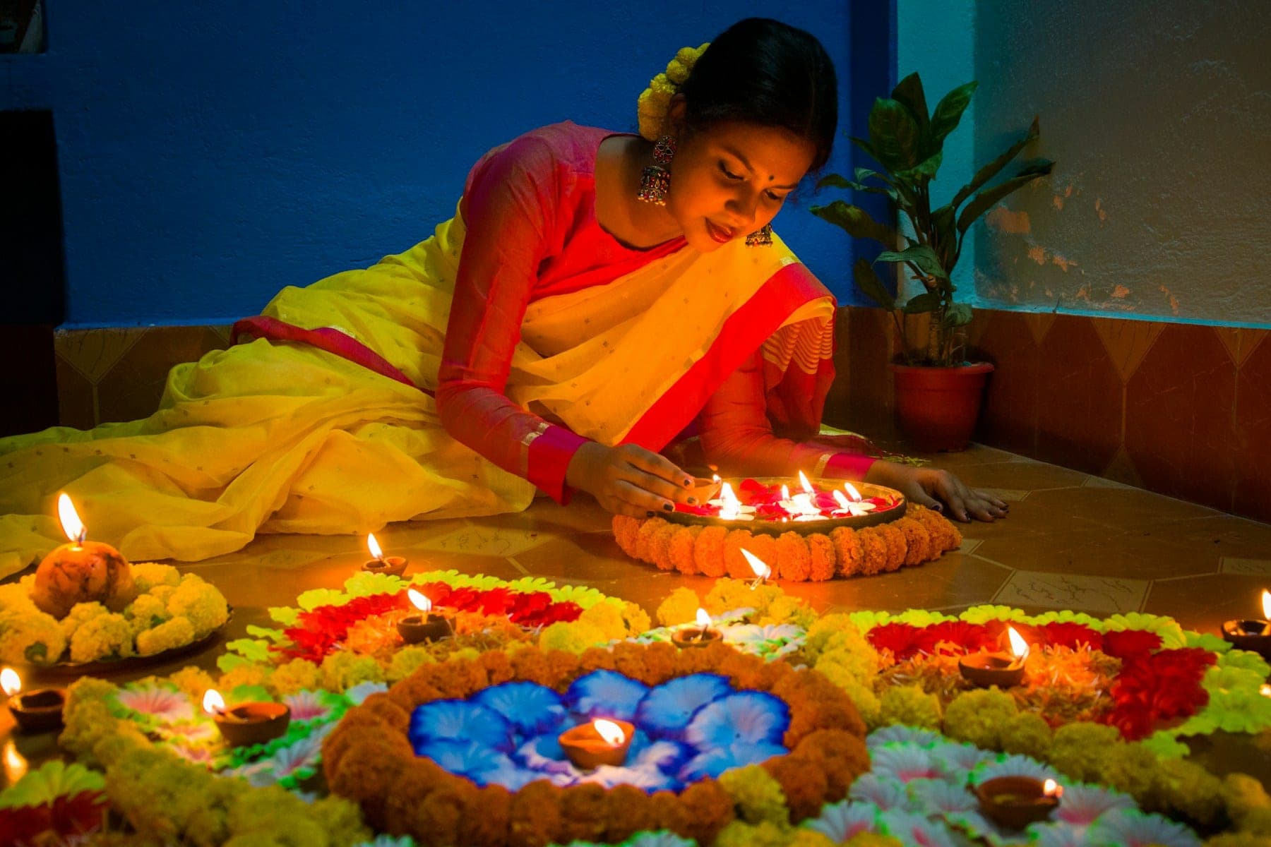 Woman in a yellow saree lighting diyas beside a flower rangoli